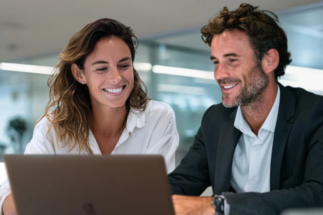 Two professionals collaborating and smiling while reviewing content on a laptop in an office.
